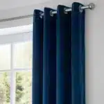 Gros plan sur la tête d'un rideau en velours bleu canard montrant les œillets métalliques argentés sur une tringle.