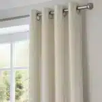 Gros plan sur un rideau en velours blanc cassé (milky white) suspendu à une tringle en métal brossé, avec des œillets. Le rideau est fermé, laissant entrevoir une fenêtre et la verdure extérieure.