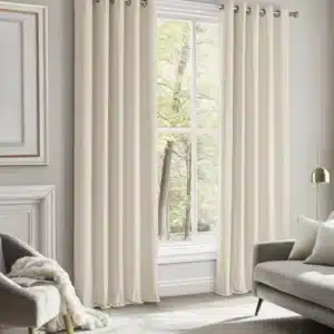 Une paire de rideaux en velours blanc cassé (milky white) habillant une grande fenêtre dans un salon moderne. La lumière naturelle éclaire la pièce, avec un fauteuil confortable et un canapé gris visibles.