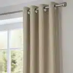 Rideau beige suspendu à une tringle métallique à œillets. Le tissu a une texture de lin légèrement striée. La scène montre une fenêtre et une partie du mur intérieur.