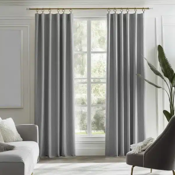 Une paire de rideaux gris moderne à texture, accrochés à une tringle décorative, encadrant une grande fenêtre dans un salon lumineux avec canapé et fauteuil.
