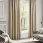 Paire de rideaux beiges, tombant au sol et encadrant une grande fenêtre de salon. Les rideaux présentent une texture de lin et sont ouverts sur une vue d'arbres. Un canapé et un fauteuil gris sont visibles dans la pièce.