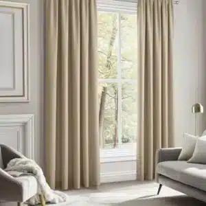 Paire de rideaux beiges, tombant au sol et encadrant une grande fenêtre de salon. Les rideaux présentent une texture de lin et sont ouverts sur une vue d'arbres. Un canapé et un fauteuil gris sont visibles dans la pièce.