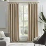 Paire de rideaux beiges avec un plissé classique, suspendus à une tringle en laiton. Le tissu a une texture de lin texturée. La scène se déroule dans un intérieur lumineux avec une fenêtre centrale, un fauteuil et une plante.