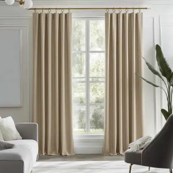 Paire de rideaux beiges avec un plissé classique, suspendus à une tringle en laiton. Le tissu a une texture de lin texturée. La scène se déroule dans un intérieur lumineux avec une fenêtre centrale, un fauteuil et une plante.