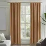 Rideaux couleur rouille ou brun noisette, plissés, suspendus à une tringle dorée dans un salon moderne avec des murs clairs, un canapé gris et une plante.
