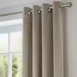 Gros plan sur des rideaux en tissu de couleur taupe ou marron clair, montrant le tissu, les plis et les œillets métalliques argentés sur une tringle.