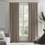 Rideaux marron clair/taupe, plissés, suspendus à une tringle dorée dans un salon moderne avec des murs clairs, un canapé gris, un fauteuil gris foncé et une vue sur la verdure.