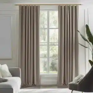 Rideaux marron clair/taupe, plissés, suspendus à une tringle dorée dans un salon moderne avec des murs clairs, un canapé gris, un fauteuil gris foncé et une vue sur la verdure.