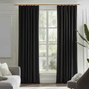 Rideaux noirs, plissés, suspendus à une tringle dorée dans un salon moderne avec des murs blancs, un canapé gris, un fauteuil gris foncé et une plante verte.