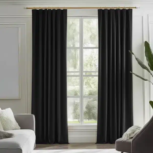 Rideaux noirs, plissés, suspendus à une tringle dorée dans un salon moderne avec des murs blancs, un canapé gris, un fauteuil gris foncé et une plante verte.