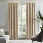 Intérieur de salon moderne avec des rideaux en velours beige remplaçant les rideaux blancs, un canapé gris clair, un fauteuil gris foncé, et une grande plante verte, créant une atmosphère chic et naturelle.