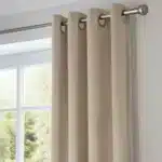 Gros plan réaliste sur la partie supérieure d'un rideau en velours beige clair, montrant la texture du tissu et les œillets métalliques argentés sur une tringle de couleur nickel brossé.